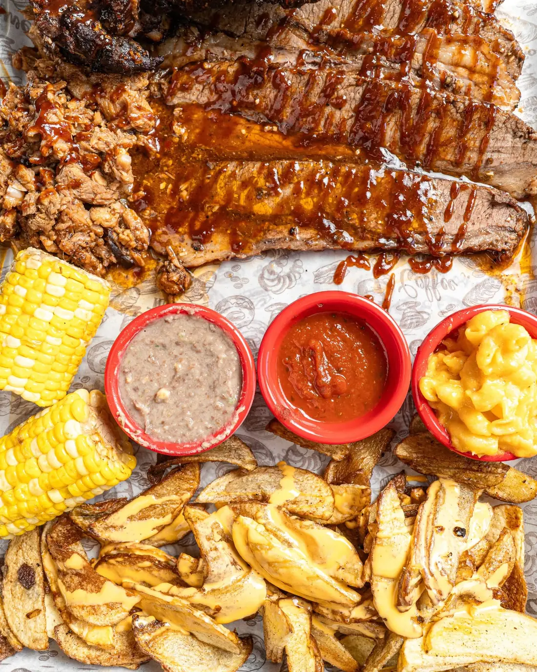 Vista cenital de una charola de brisket, costillas, pulled pork, elote, papas y salsas